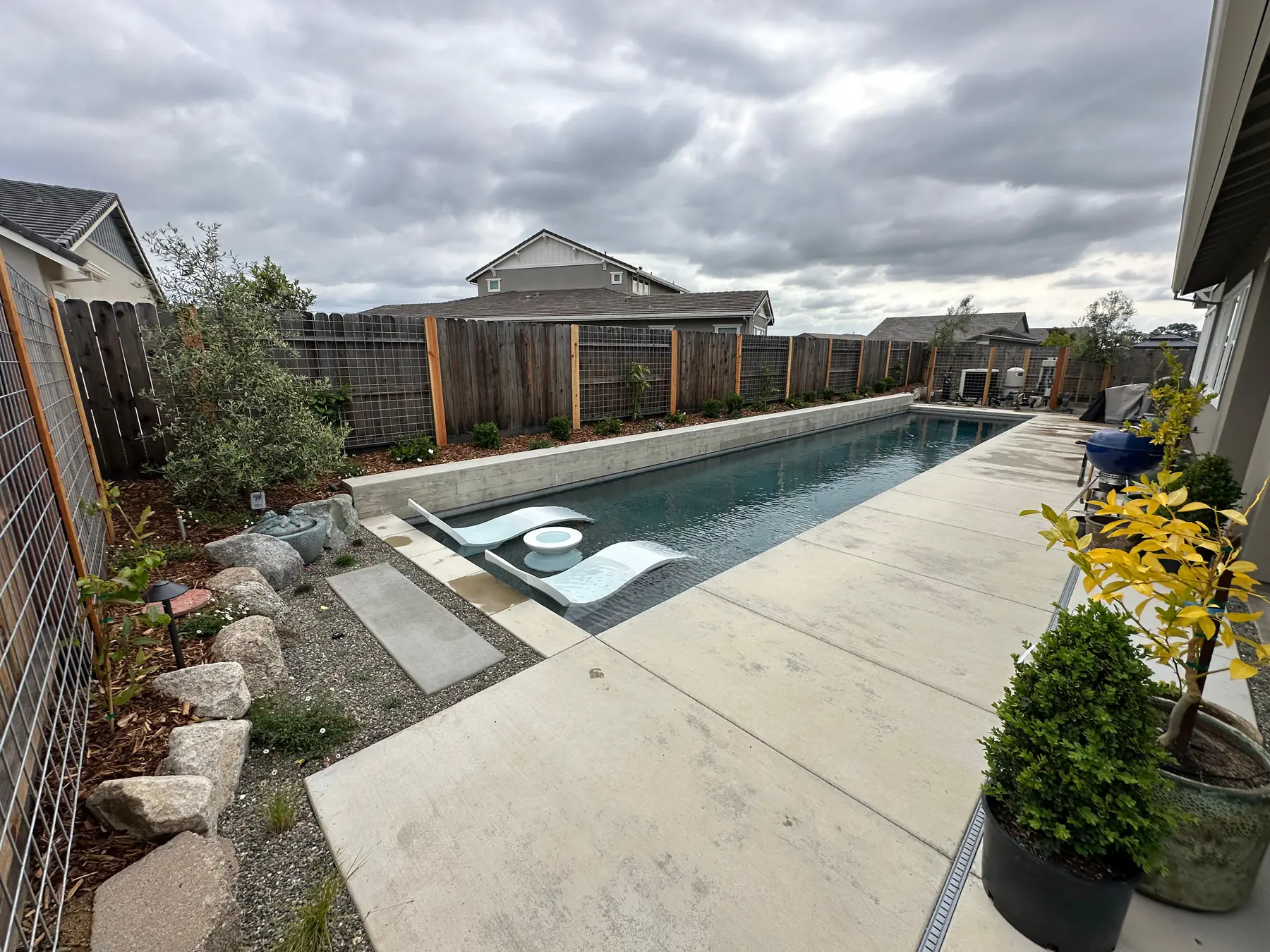 Poolside landscape with concrete patio, planting, boulders, and modern fence